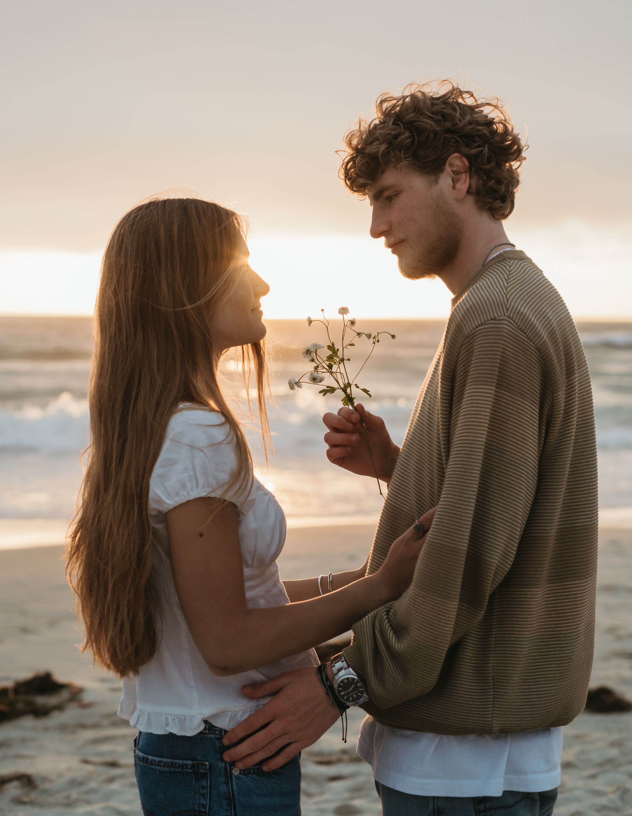 On a sunset beach, a romantic couple embraces as they share a tender moment with a small flower at golden hour.