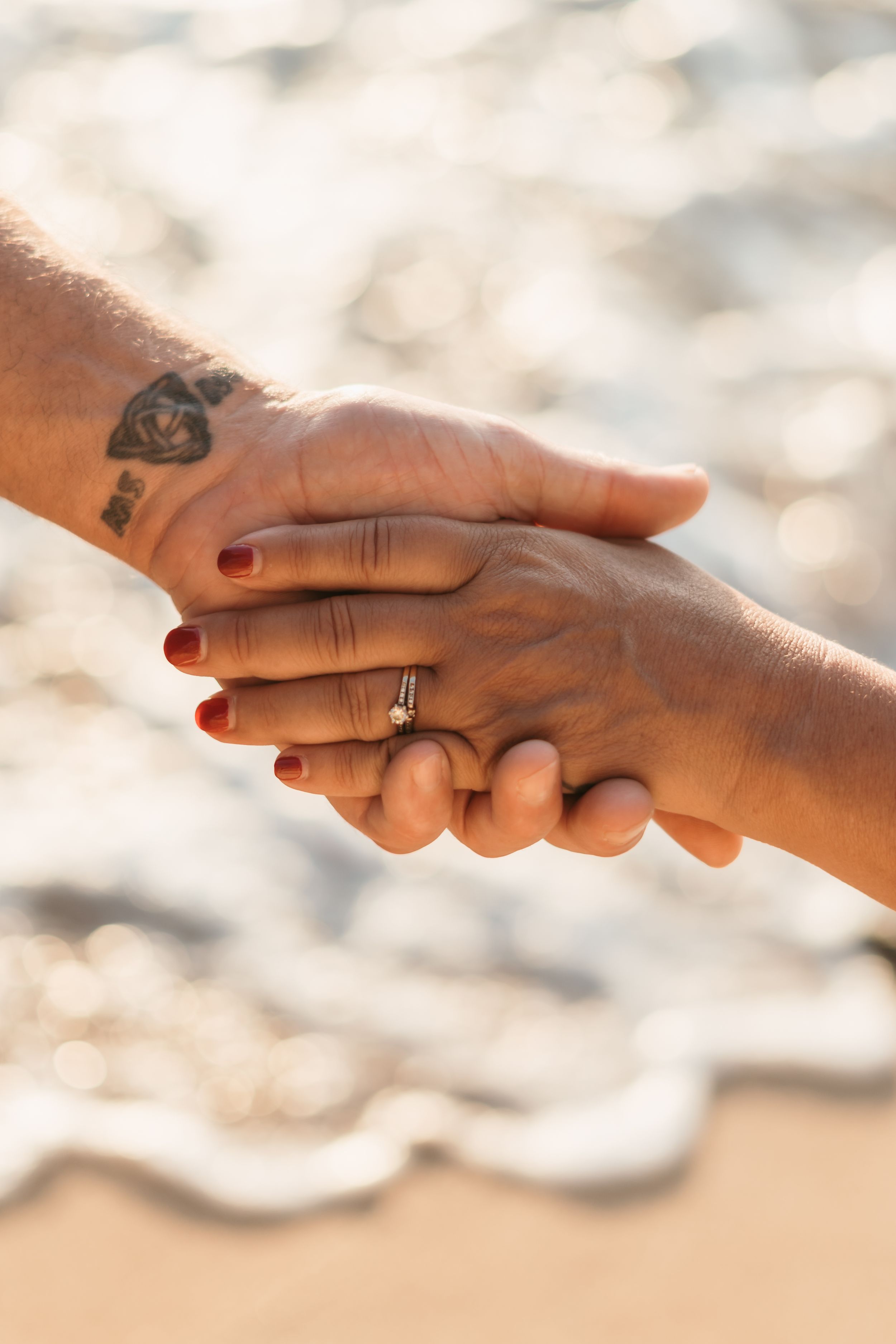 Close up of two hands gently clasping together with a wedding ring visible on a blurred beach background.