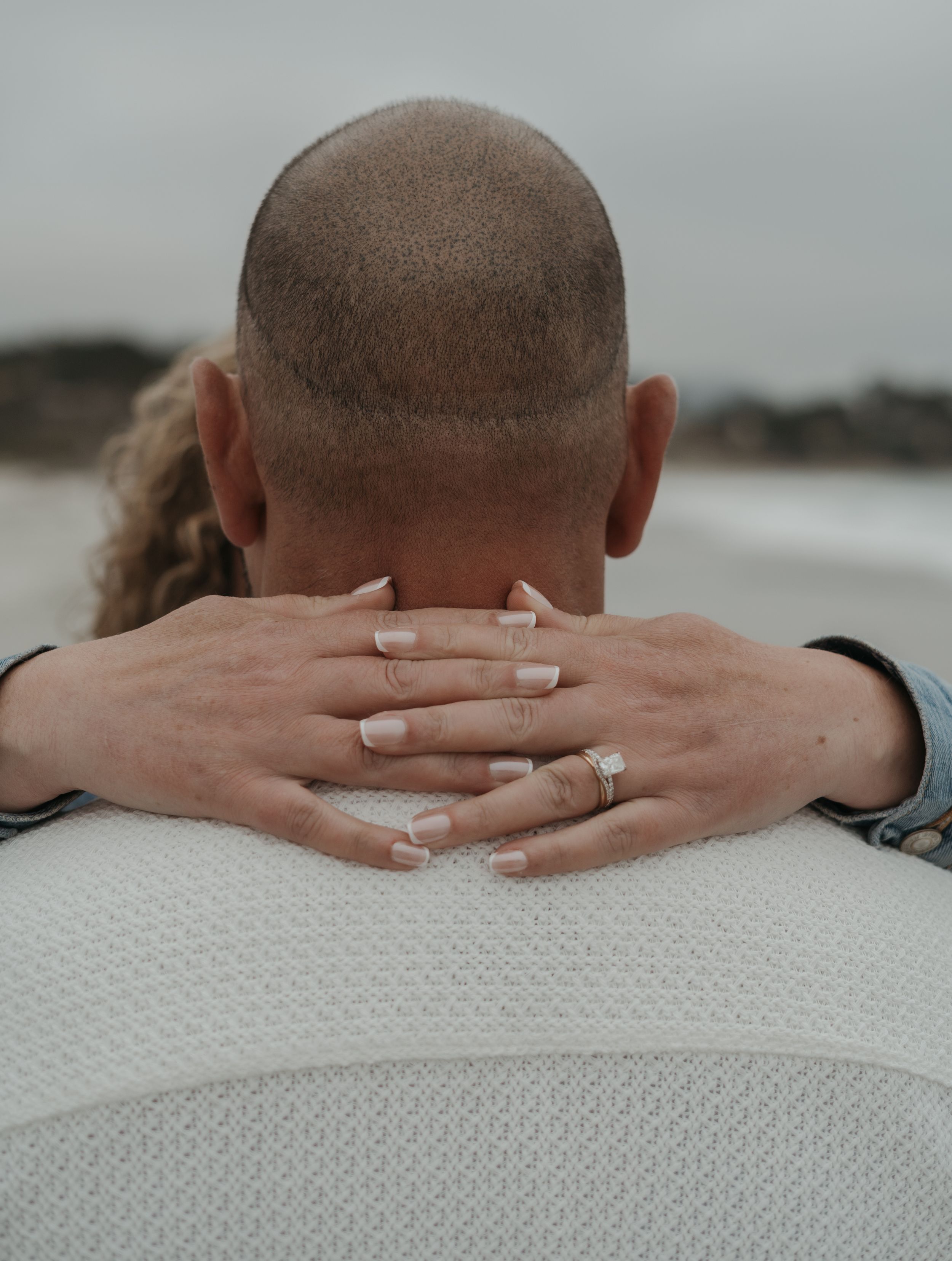 A close up view of hands clasped behind a head against a light colored background, showing an engagement ring.