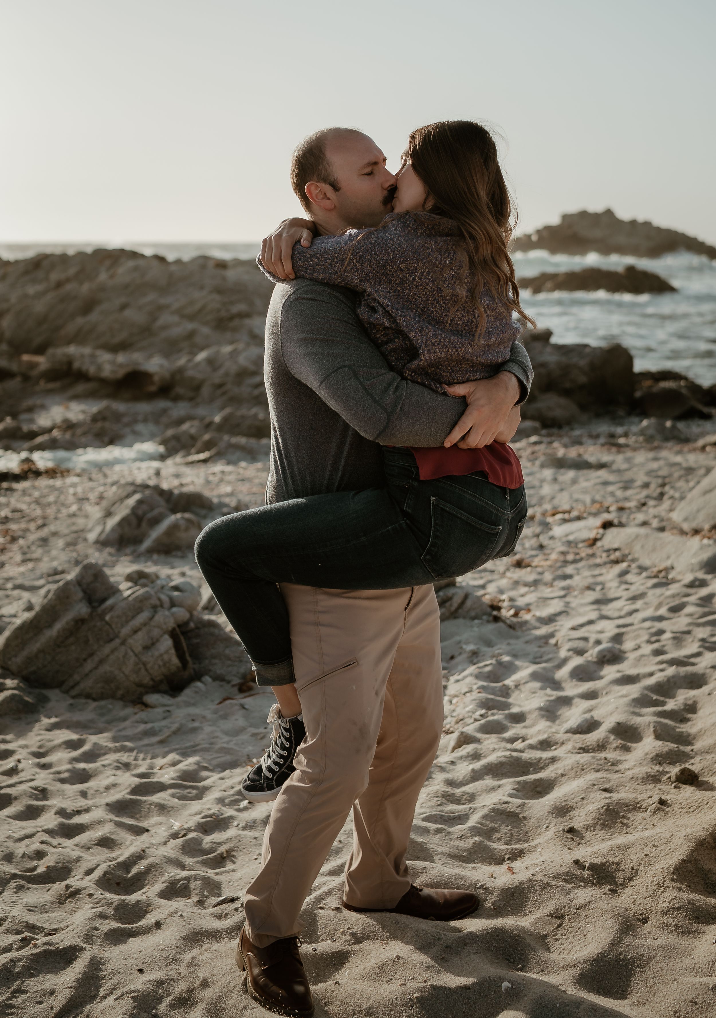 A couple embraces while sharing an intimate moment on a rocky beach at sunset with ocean waves in the background.