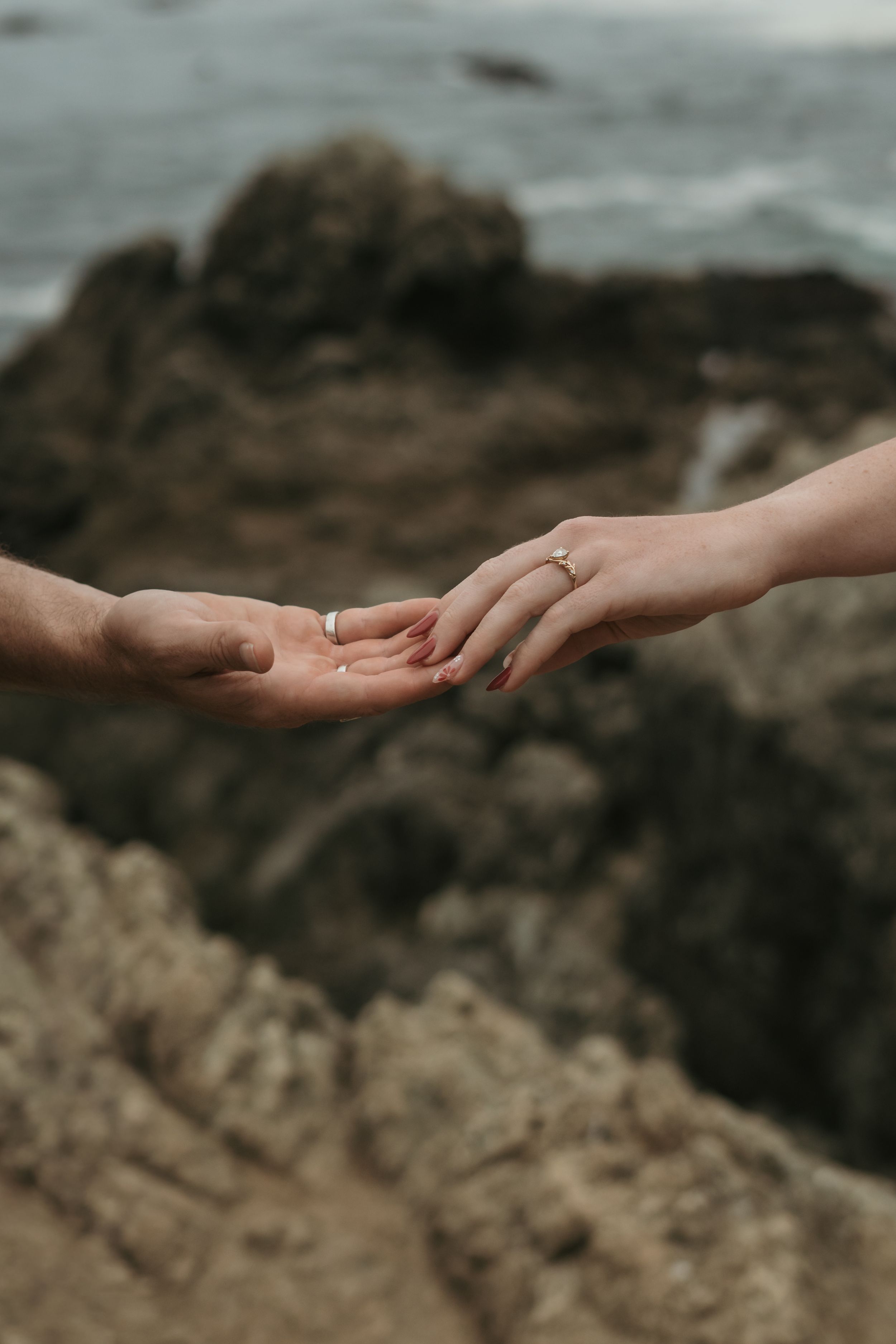 Two hands reaching towards each other against a blurred rocky coastal background in a romantic gesture.