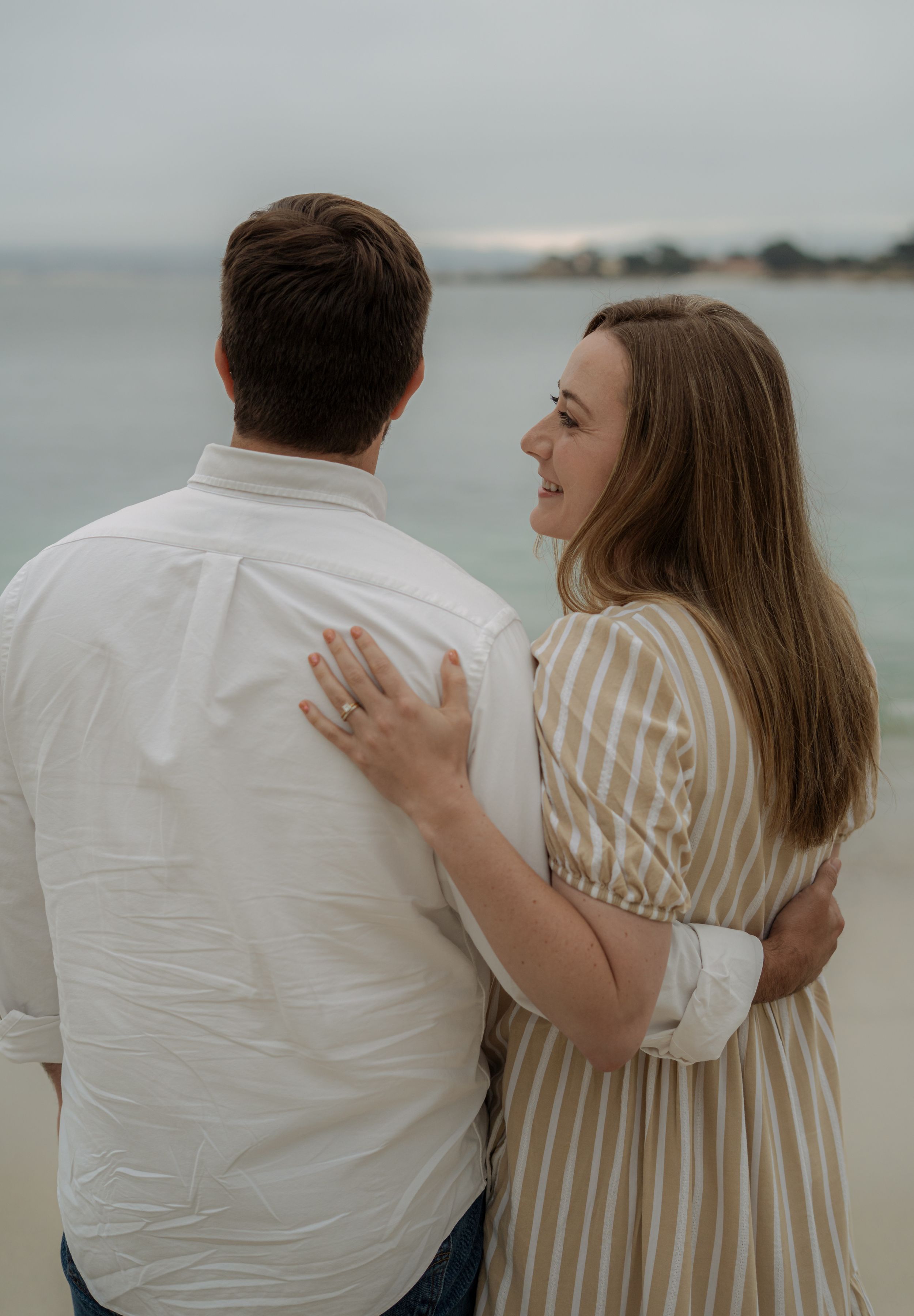 A couple embraces on a cloudy beach wearing white and striped clothing, captured in a romantic photo series.