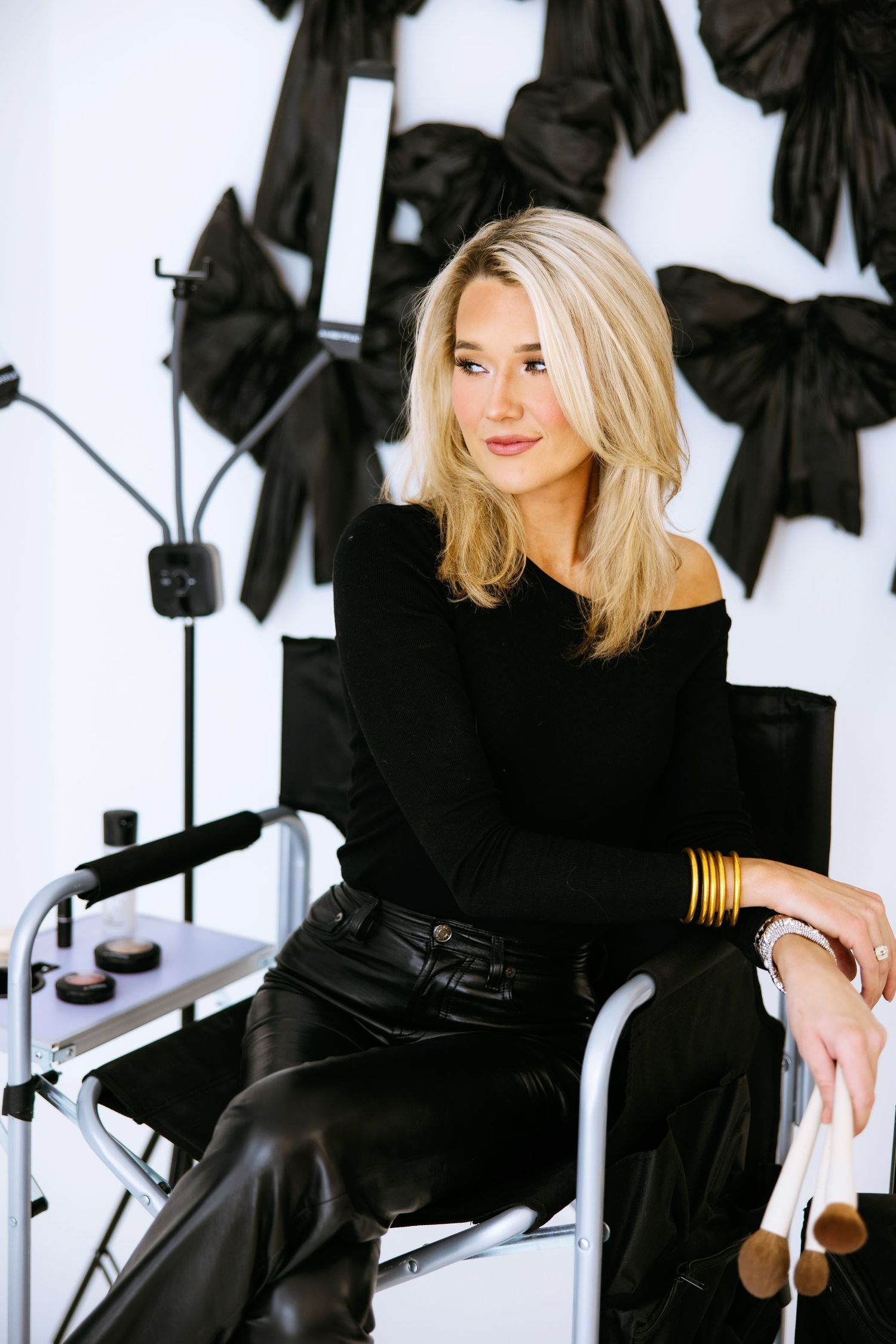 Portrait of person in all-black outfit with gold accents sitting in studio with lighting equipment visible in background.