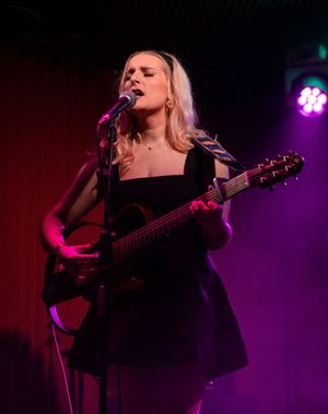 Singer-songwriter performing with guitar on dimly lit stage with purple ambient lighting.