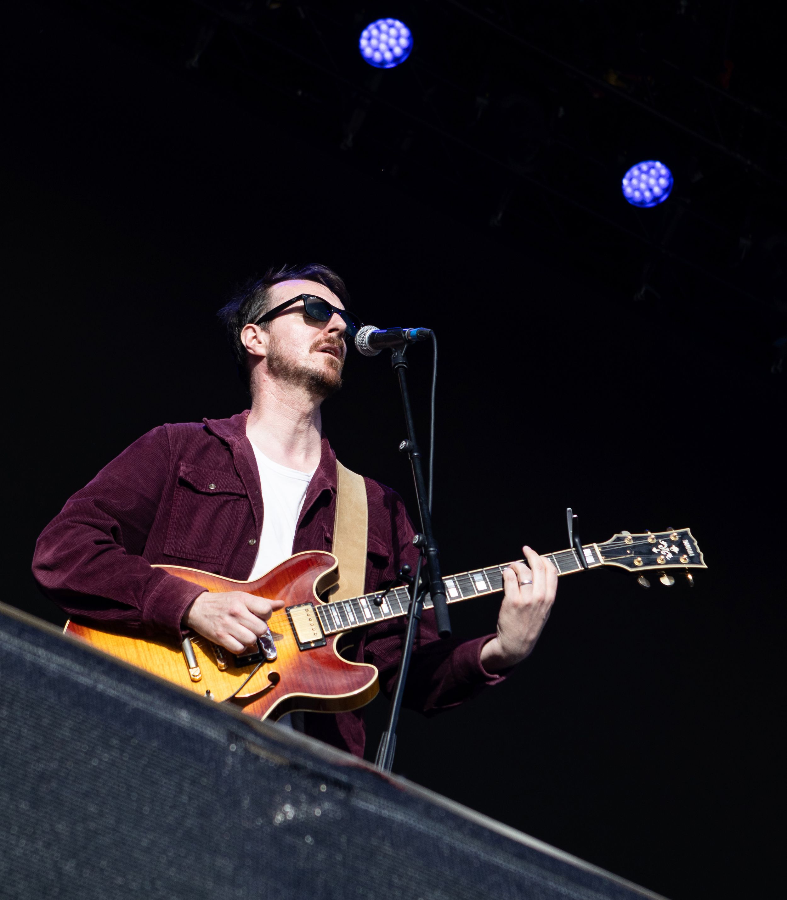 Male guitarist in sunglasses playing a semi-hollow electric guitar on stage under blue lights.