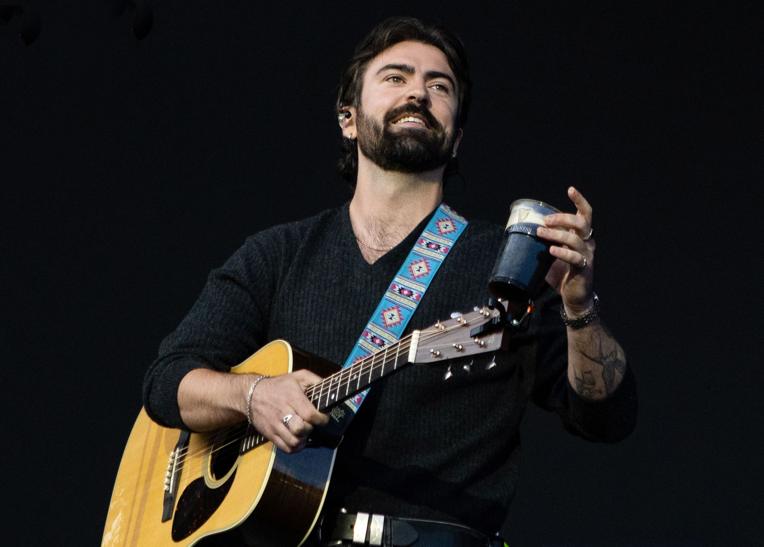 Bearded male musician playing acoustic guitar on stage with decorative strap, holding a small cup while performing live.