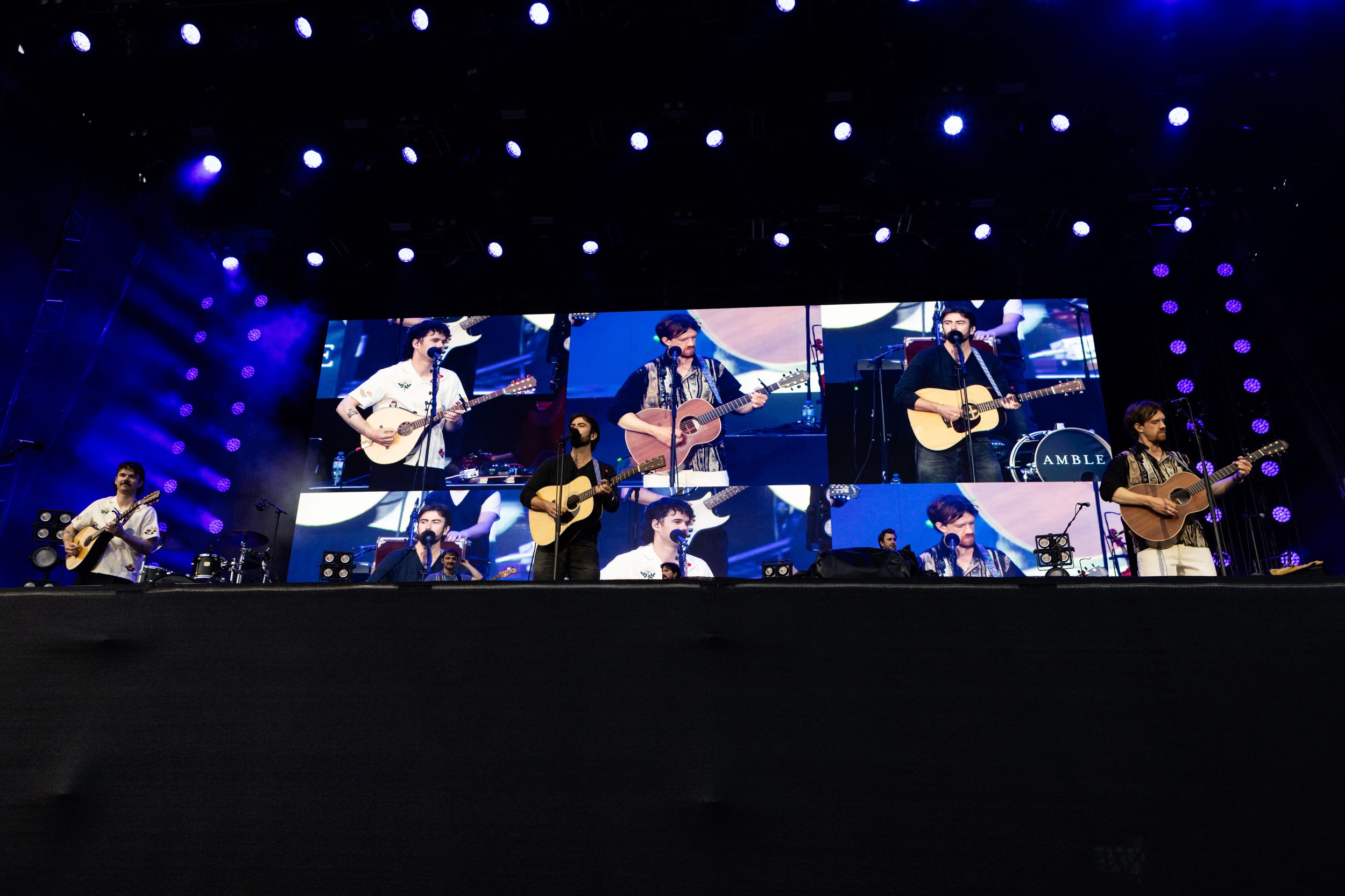 Band performing on stage with large video screen backdrop at outdoor concert.