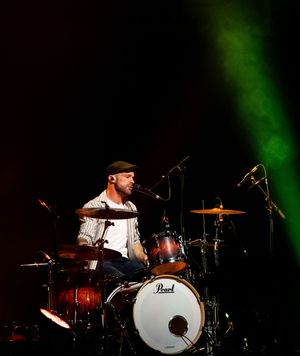 Drummer performing on a Pearl drum kit illuminated by green stage lighting during a live concert performance.
