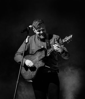 A musician performs on stage with an acoustic guitar in dramatic black and white lighting with fog effects in the background.