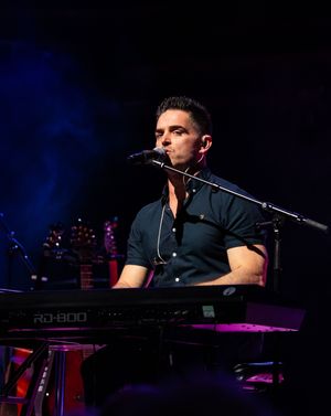 A musician performs at a piano keyboard on a dark stage with dramatic blue lighting in the background.