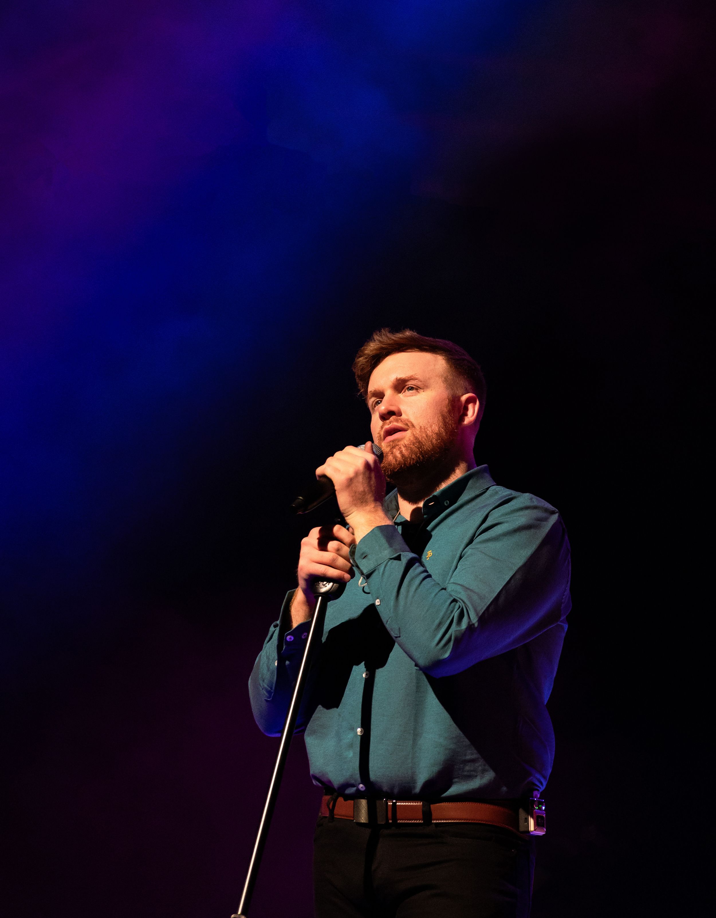 A performer in a green button-up shirt sings into a microphone on stage with blue lighting in the background.
