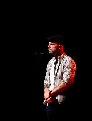 A performer in a striped shirt and dark beret stands in dramatic stage lighting against a black background.