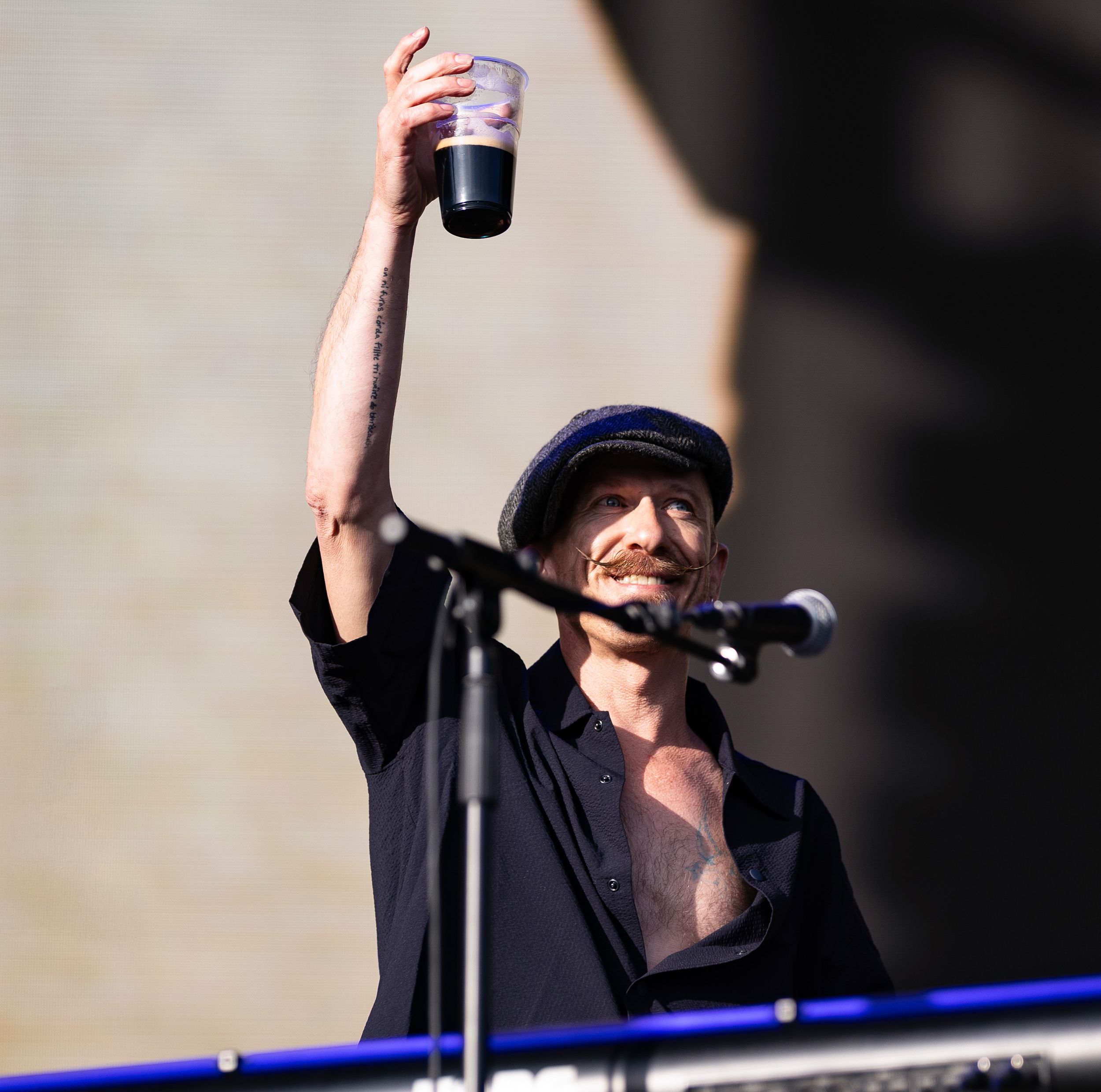 Man in black shirt and hat raises a cup triumphantly while standing at a microphone on an outdoor stage.