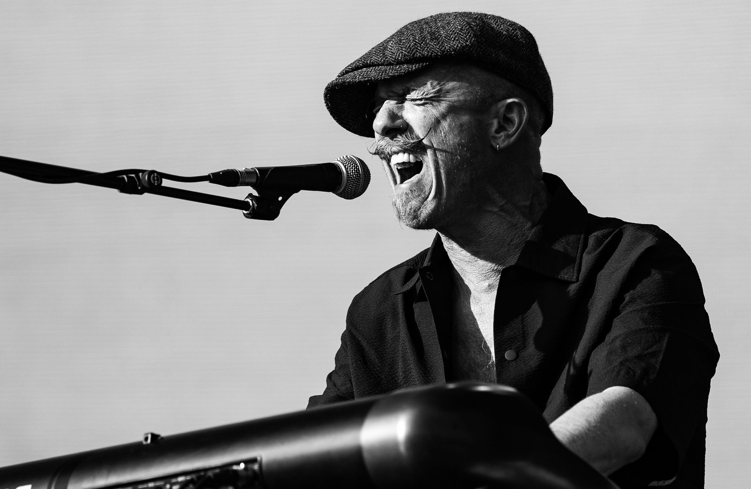 A bald man in a flat cap sings passionately into a microphone at a keyboard in black and white.