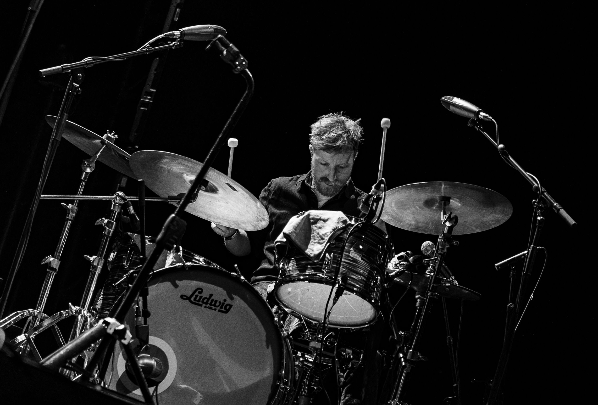 Dramatic black and white shot of drummer performing behind drum kit on dark stage.