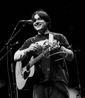 Performer smiles while playing acoustic guitar on stage in black and white photo.
