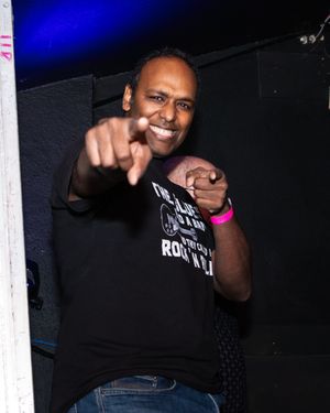 Performer in a black t-shirt points and smiles energetically at a concert venue with blue lighting.
