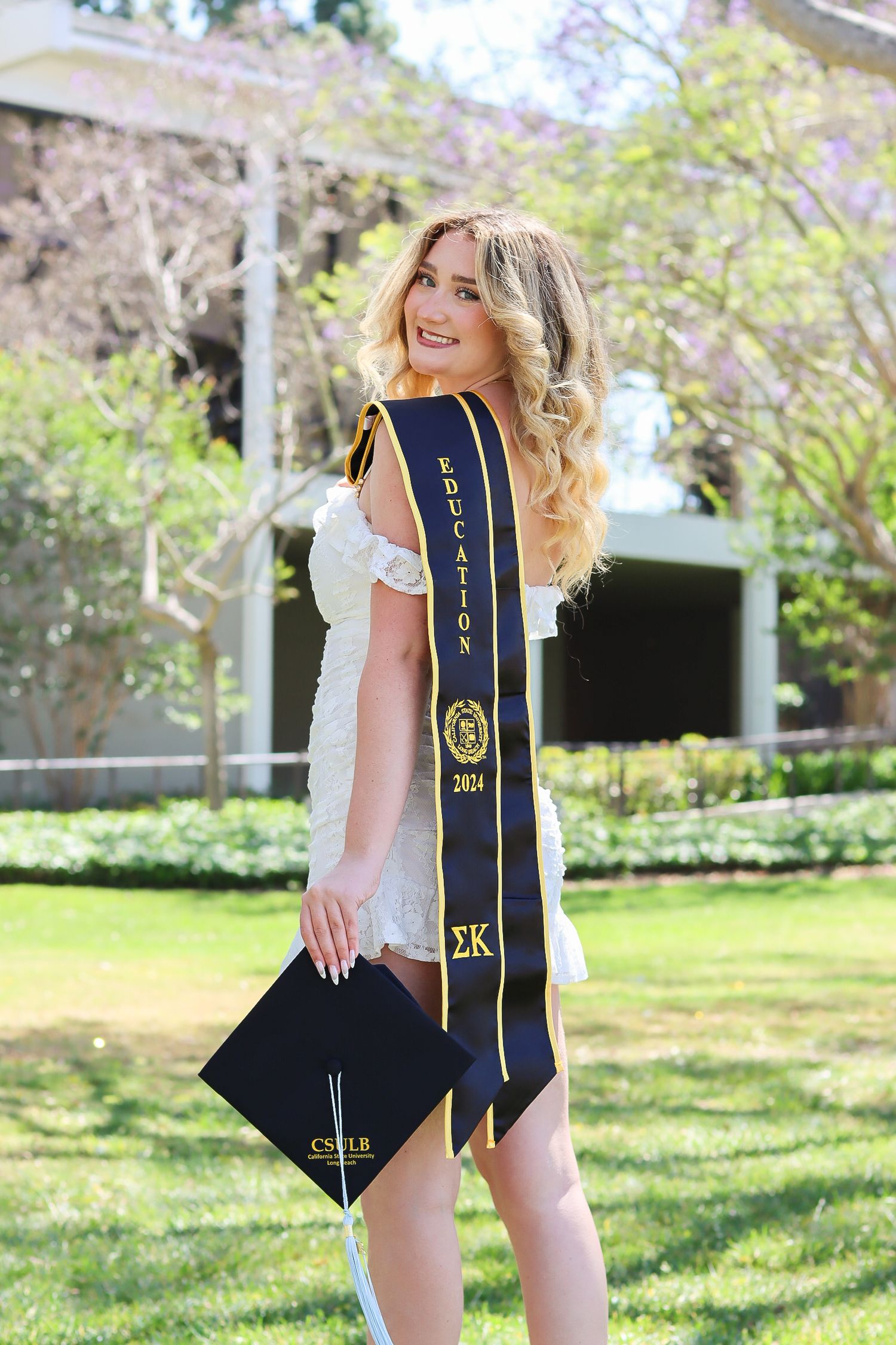 Graduate in white dress poses with graduation sash and cap on sunny grass lawn.