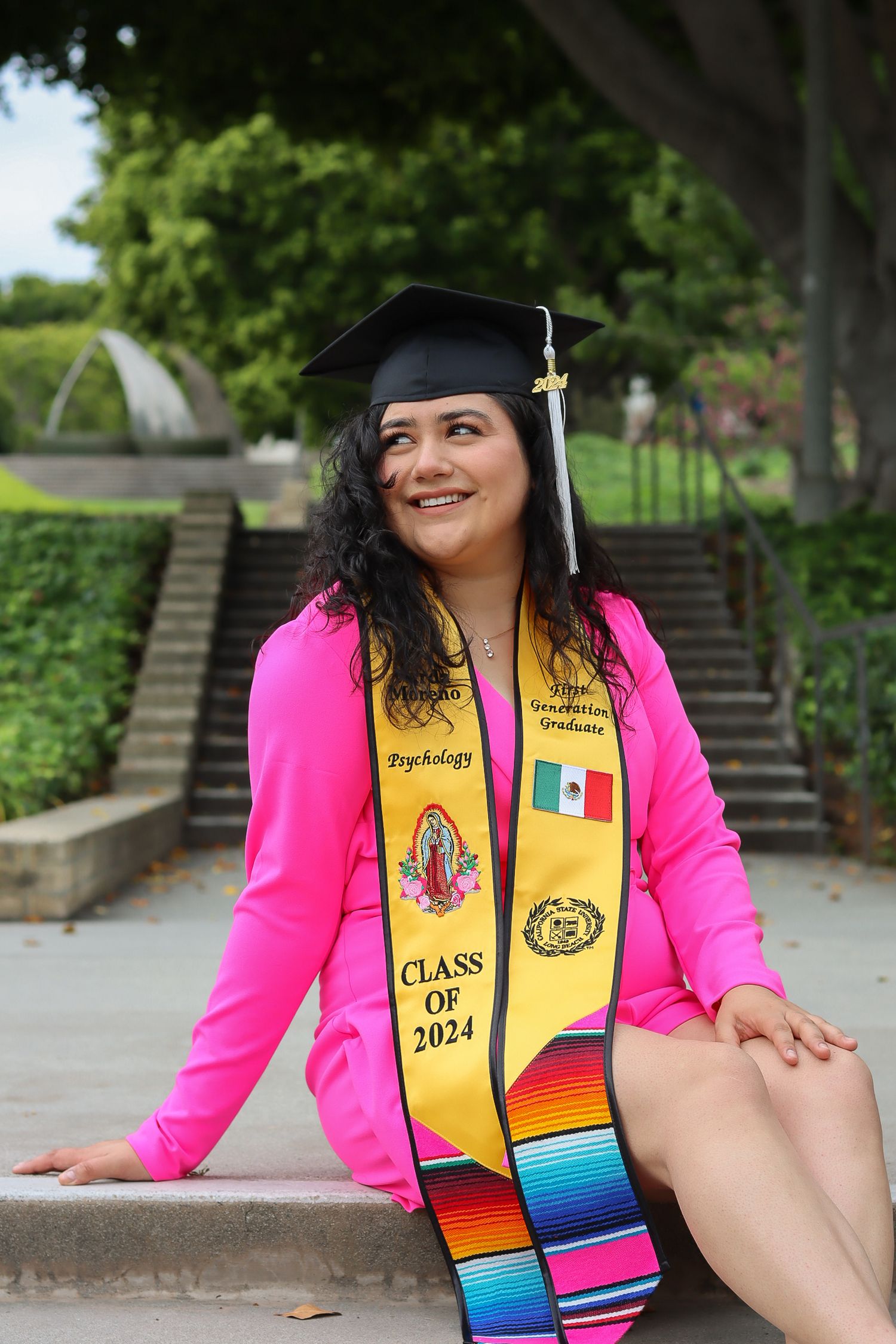 Graduate in pink blazer and decorated stole poses on campus steps with Mexican flag sash.