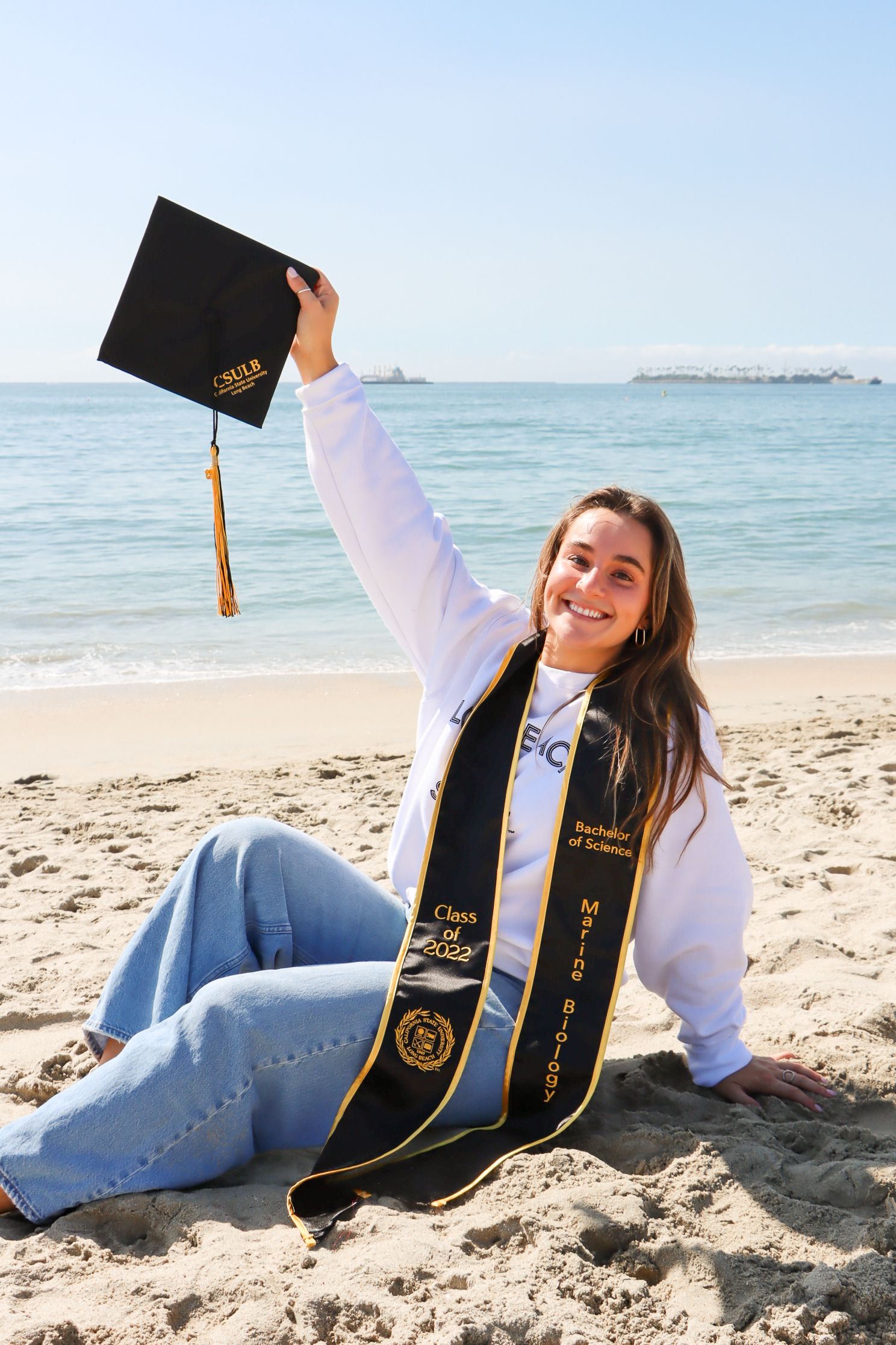 Graduate in white sweater and graduation stole poses on beach with cap and ocean view.