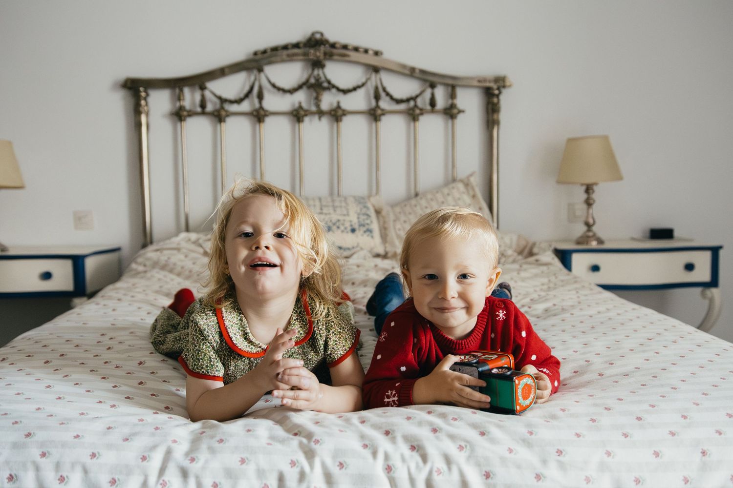 Two children wearing holiday sweaters lay together laughing on a bed with an ornate metal headboard.