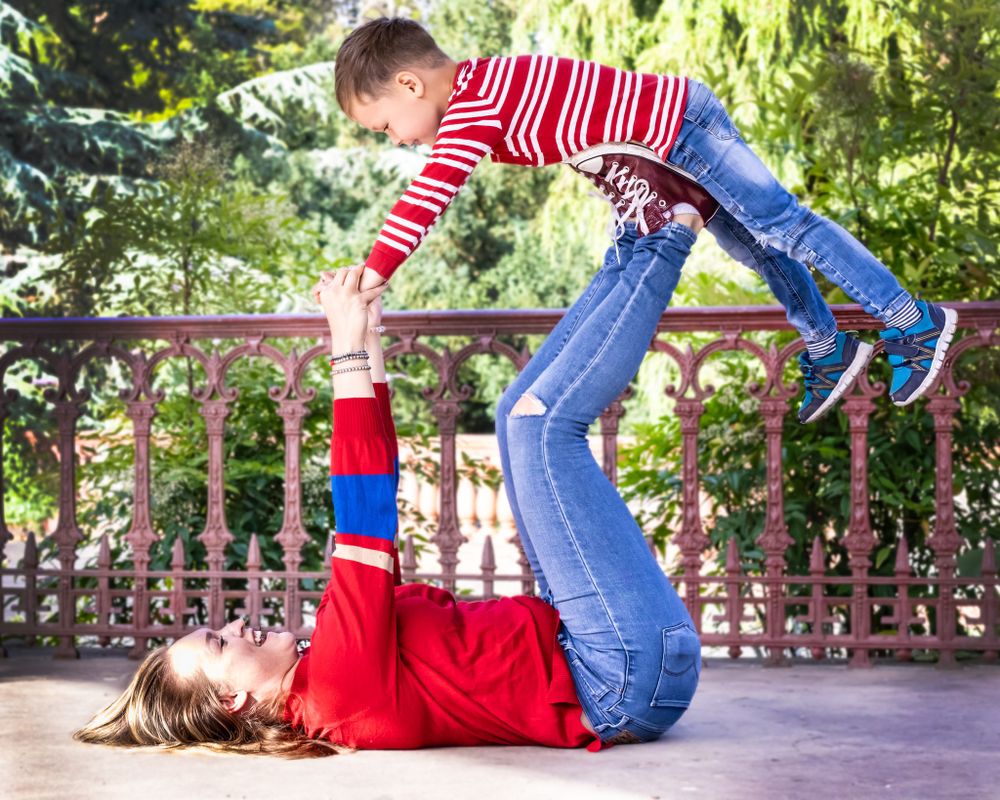 Playful family moment with parent and child doing airplane pose on outdoor patio.