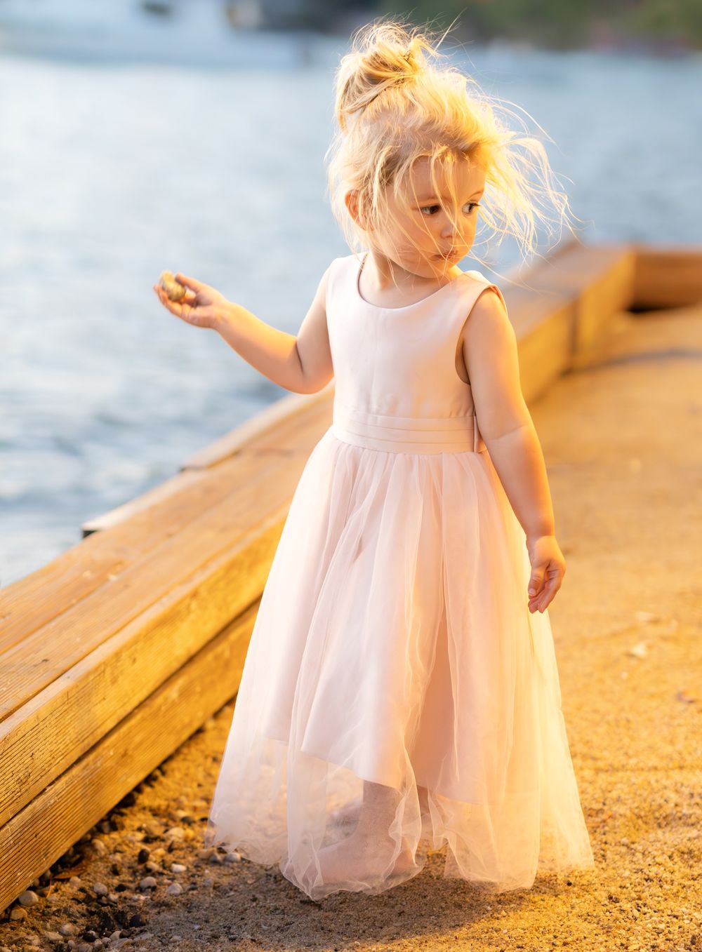 Young child in white dress standing on wooden dock at sunset.