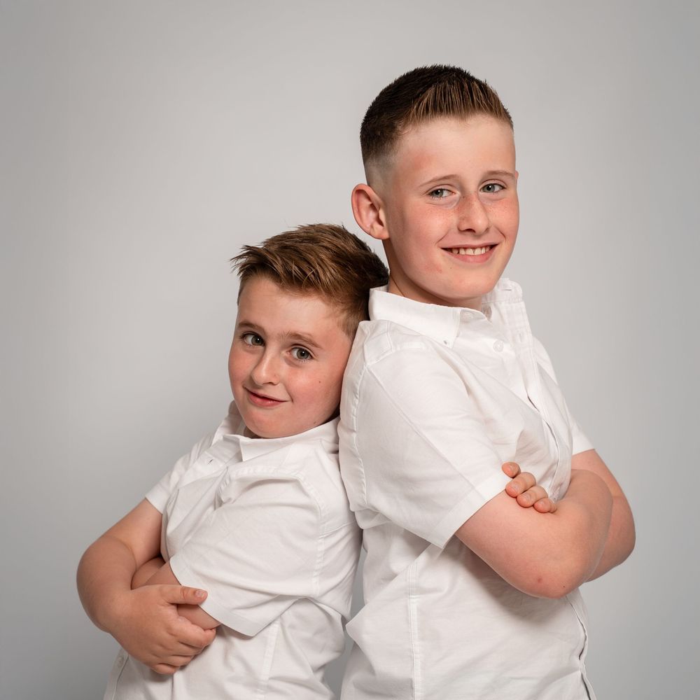 Two young children stand back-to-back in white shirts against a grey background, posing with crossed arms and smiling.