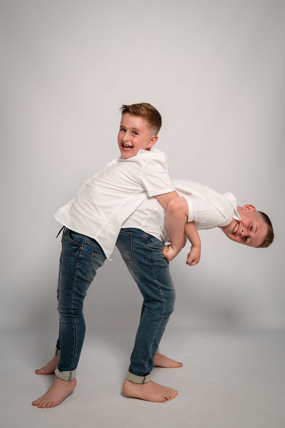 Two young males in white shirts and jeans pose playfully while barefoot against a gray studio backdrop.