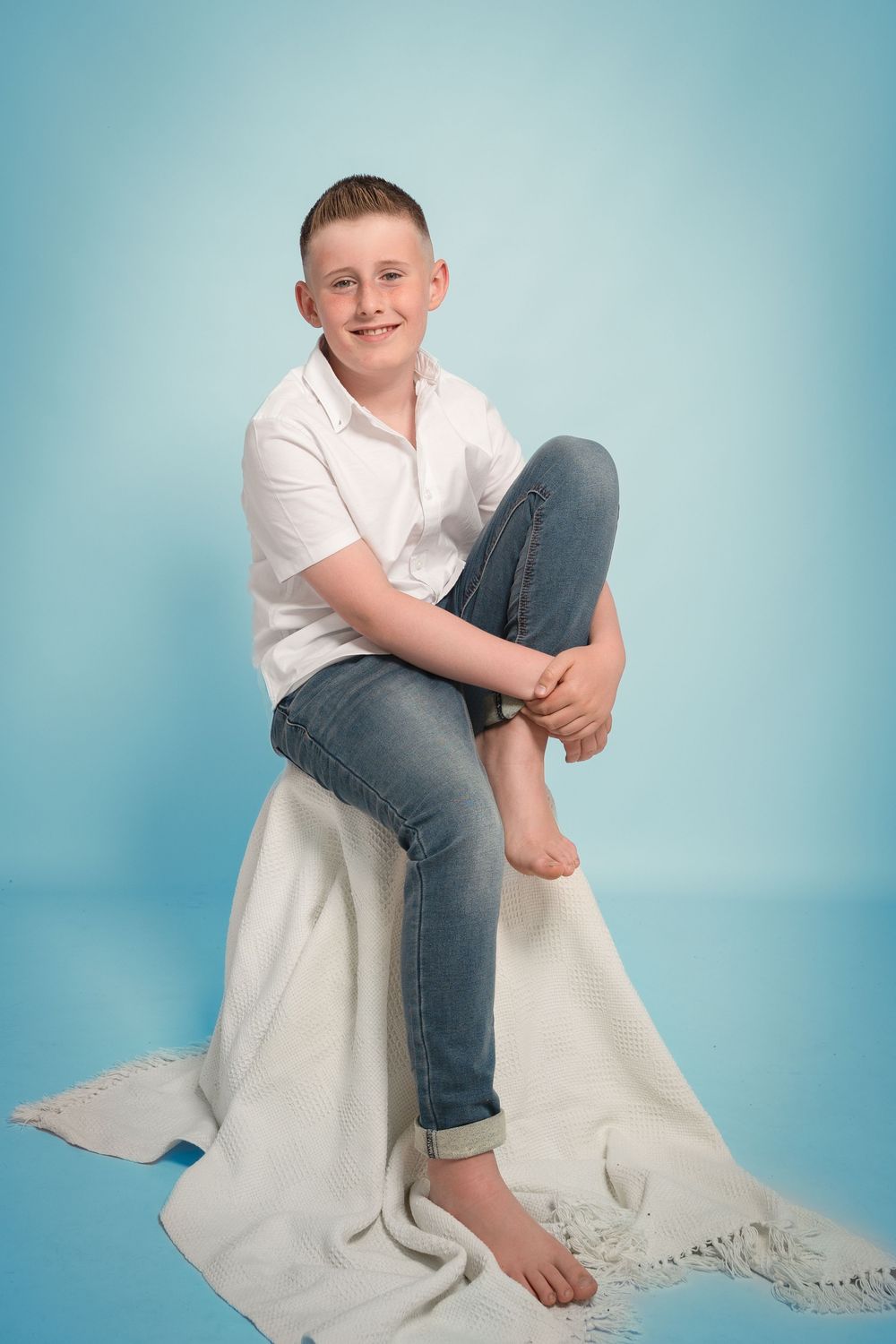 A person in a white shirt and jeans sits barefoot on a white blanket against a blue background for a studio portrait.