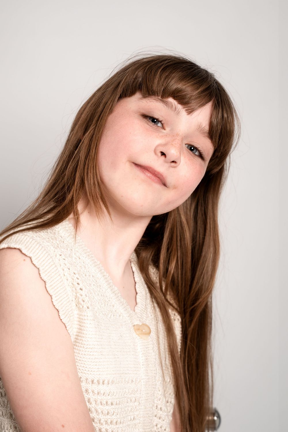 Portrait of someone with long brown hair wearing a cream-colored lace top against a light background, tilting head and smiling.