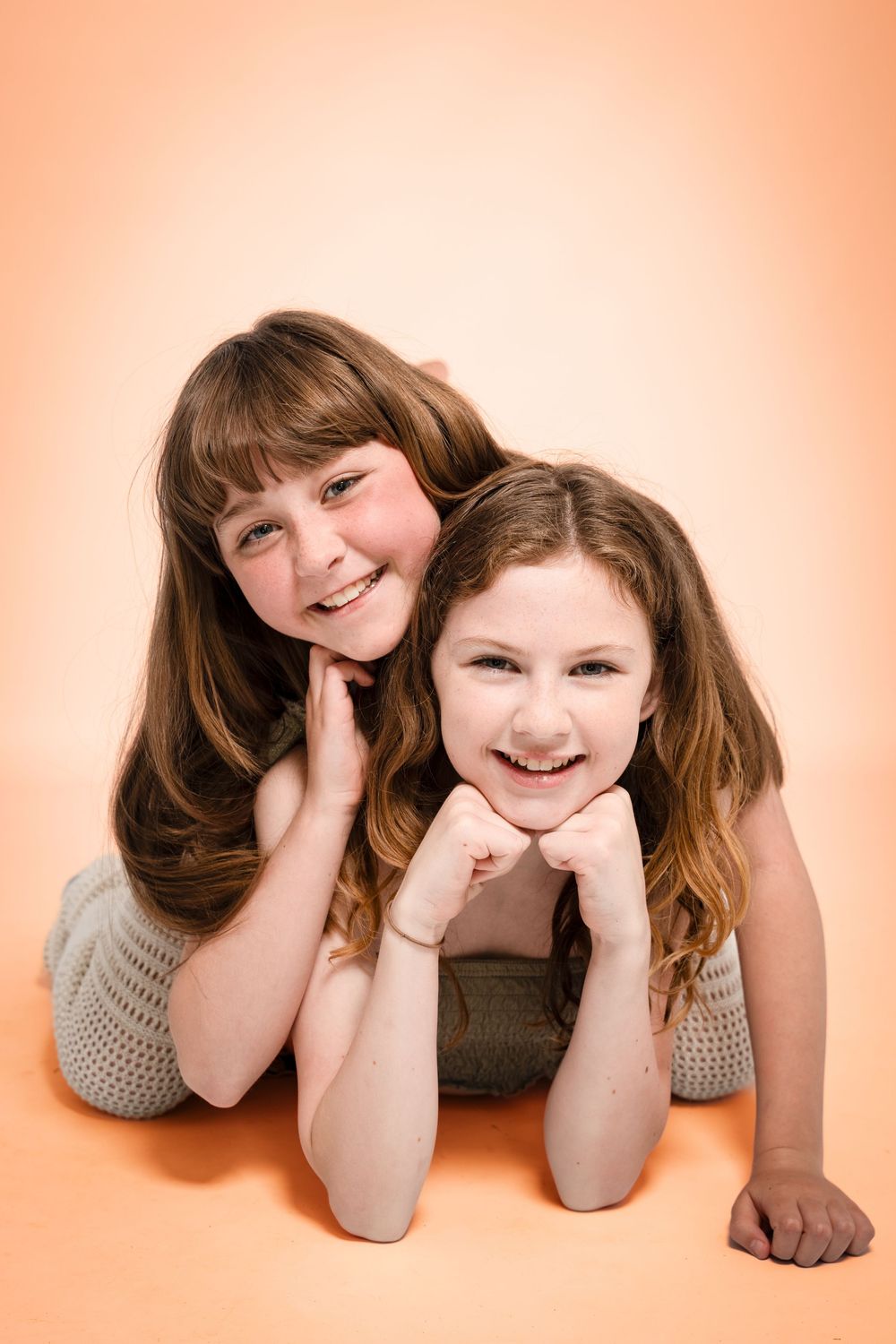 Two young people lying on a peachy-orange background, smiling warmly while posing together in casual outfits.