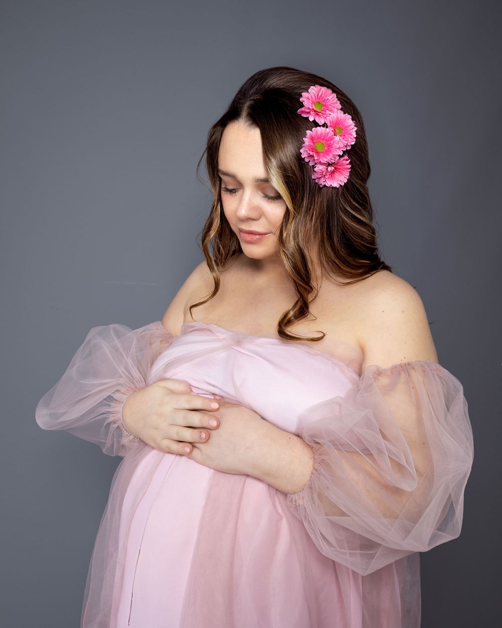 Soft maternity portrait in pink tulle dress with orchid hair flowers against gray background.