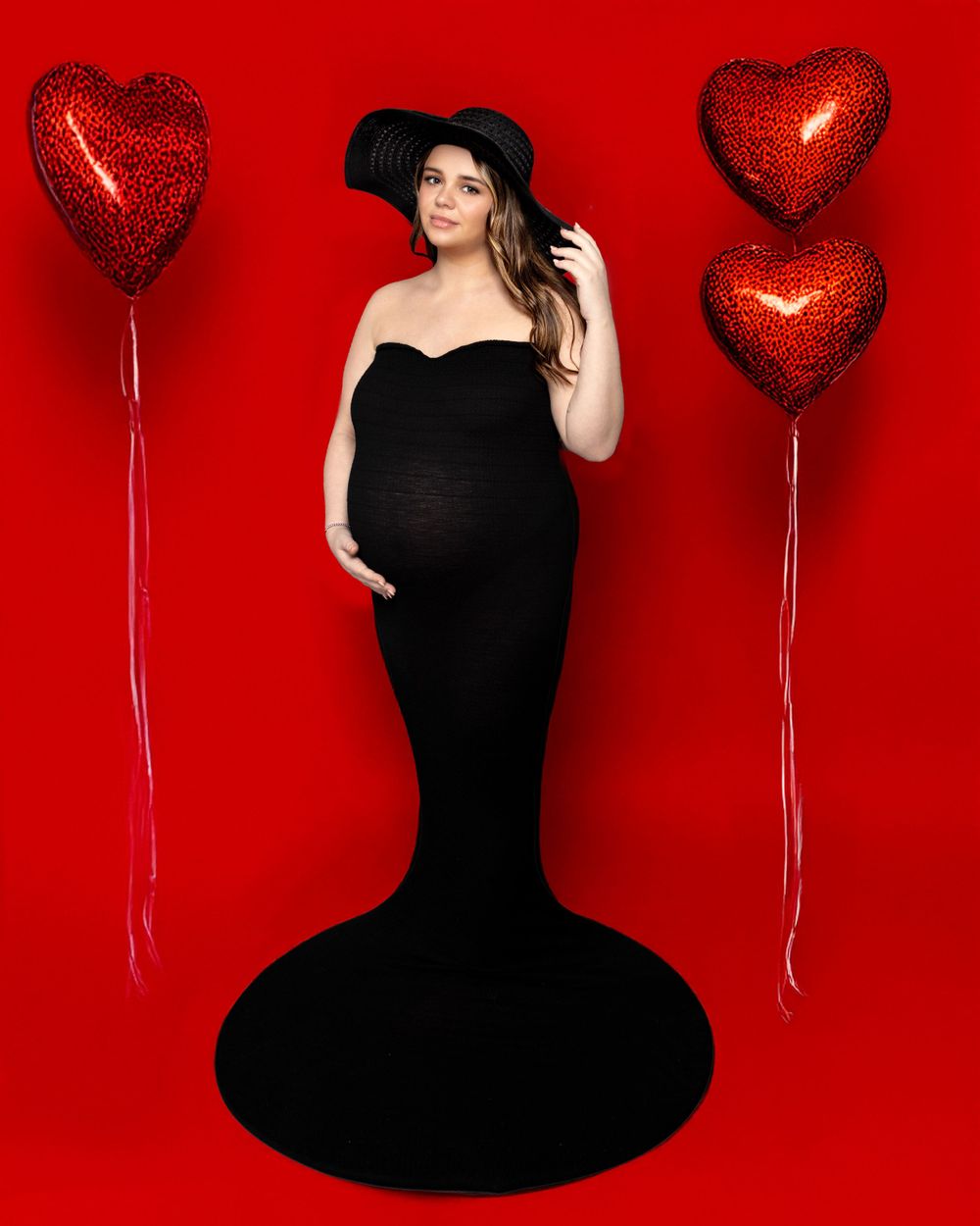 Dramatic maternity photo with black mermaid gown and red heart balloons against scarlet backdrop.