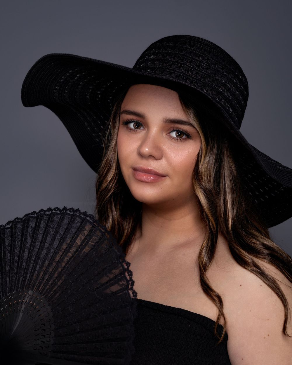 Glamour portrait featuring a wide-brim black hat and matching fan against dark background.
