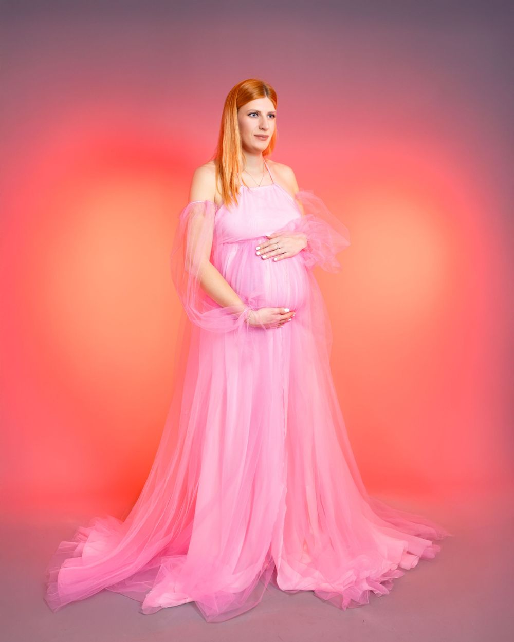Dreamy maternity portrait in pink tulle gown against coral-colored background.