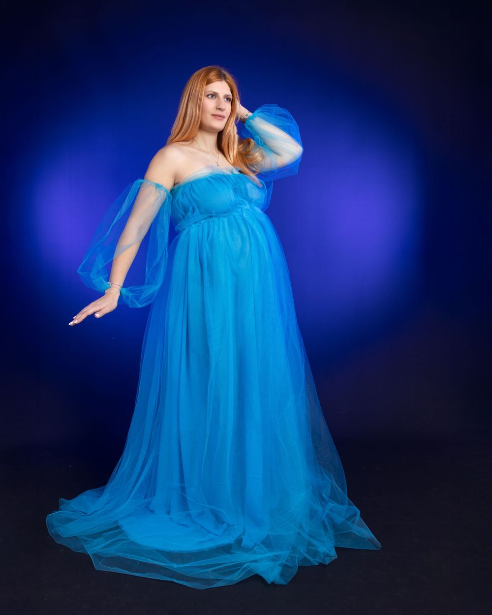 Flowing blue tulle maternity gown portrait against deep blue illuminated background.
