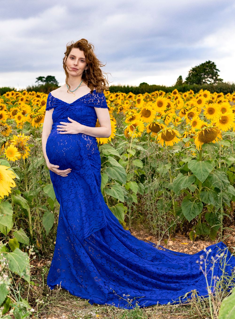 Stunning maternity photo in a royal blue lace gown with trailing train in a sunflower field.