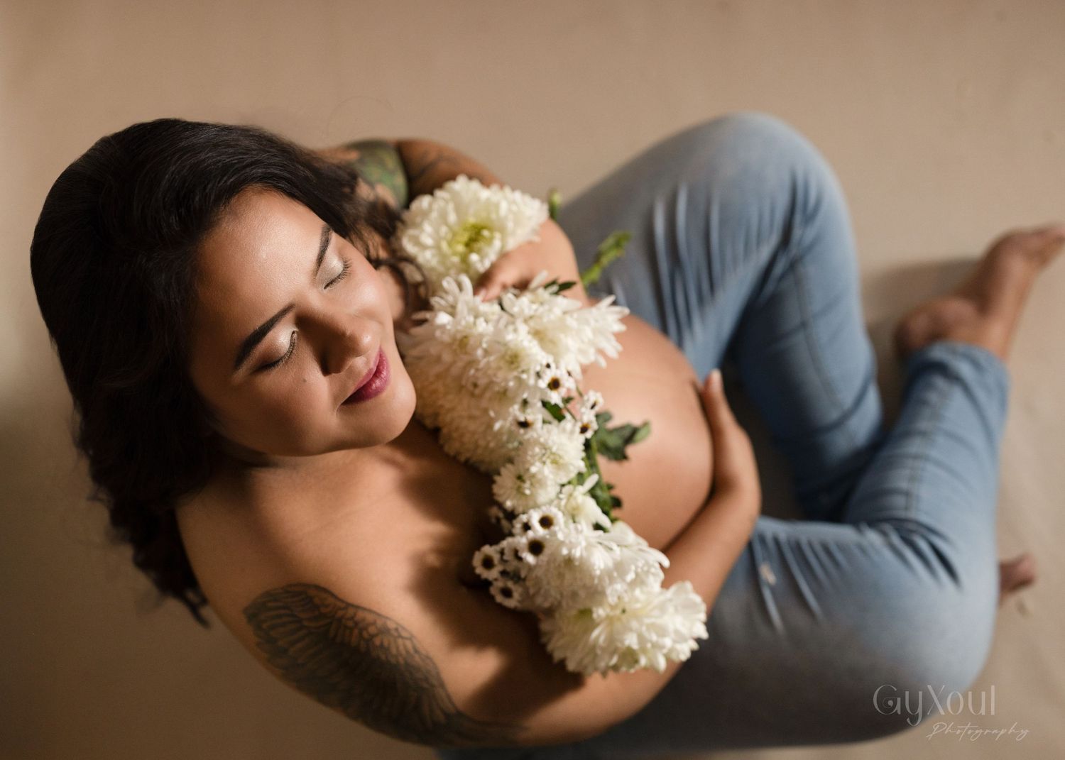 Artistic maternity photo with white flowers and relaxed pose on beige backdrop.