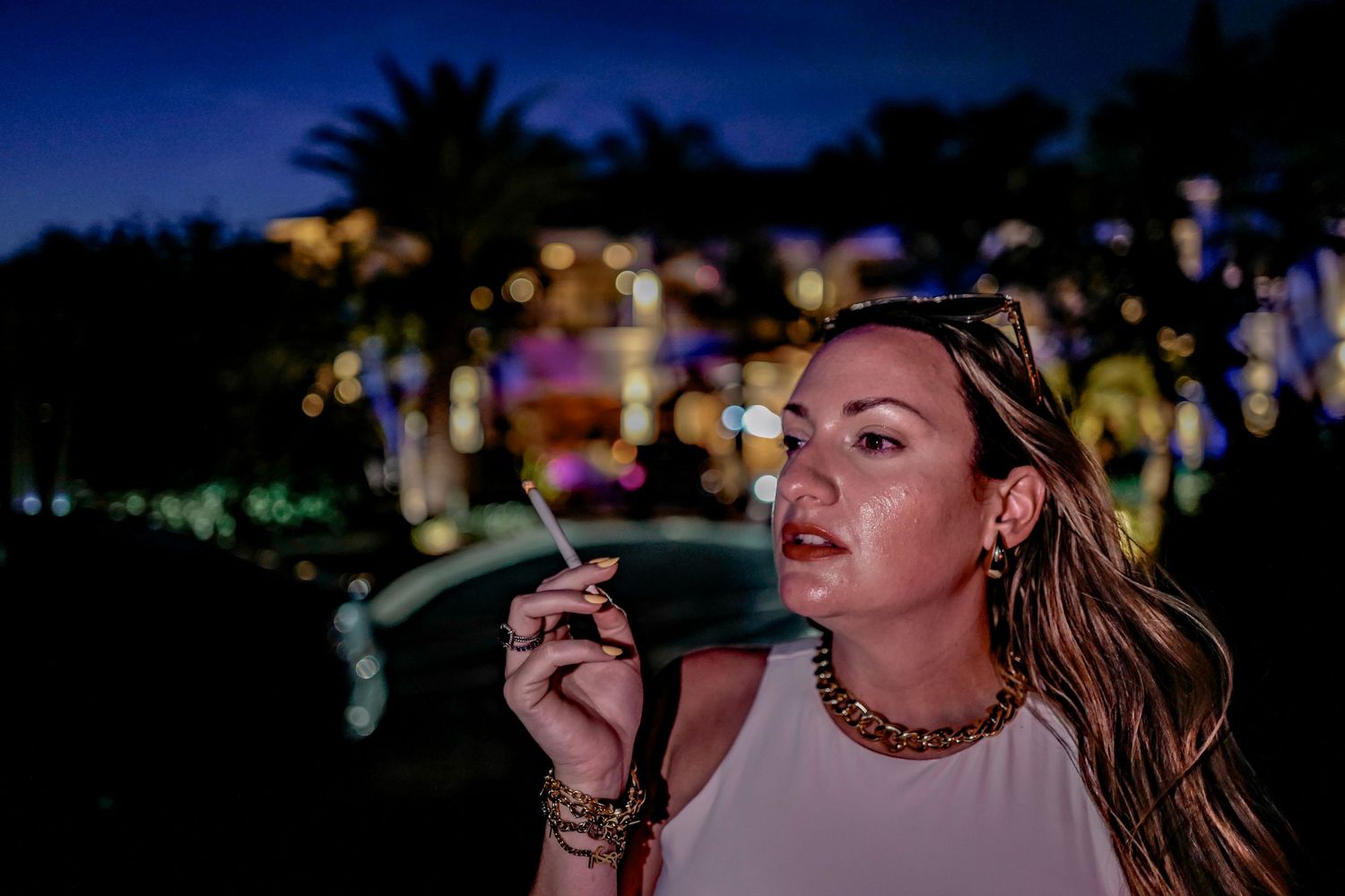 Someone smoking a cigarette outdoors at night with colorful lights and palm trees in the dark background.