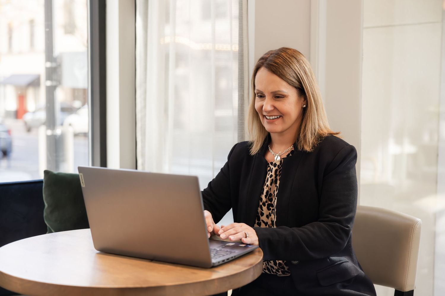 Professional working on laptop at round table by window in casual business attire.