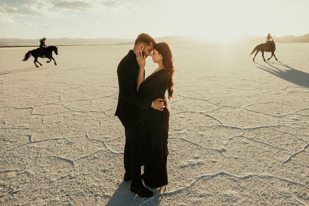 Two people embrace on a dry cracked desert with horse riders silhouetted in the background during sunset.