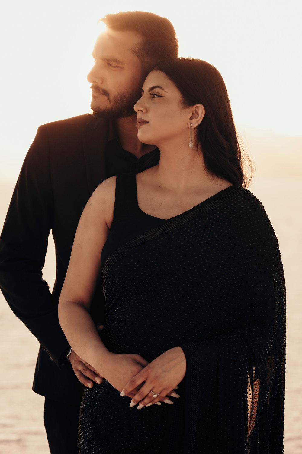 A couple embraces in a romantic sunset silhouette, with the woman wearing an elegant black evening dress and earrings.