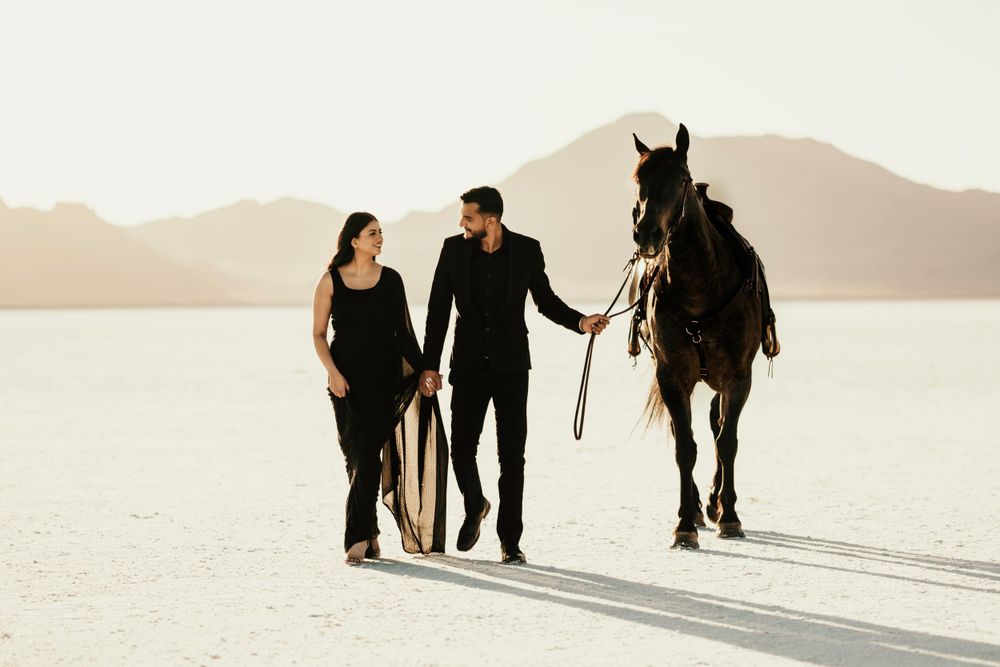 Couple walking together on white salt flats with their dark horse during sunset in a peaceful desert setting.