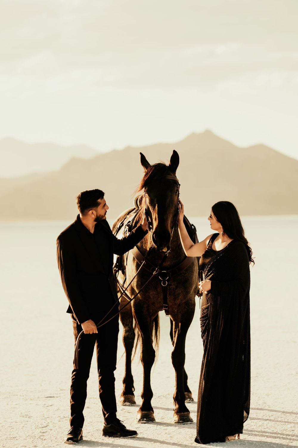 A couple stands with their horse on a salt flat at sunset, silhouetted against mountains in the background.