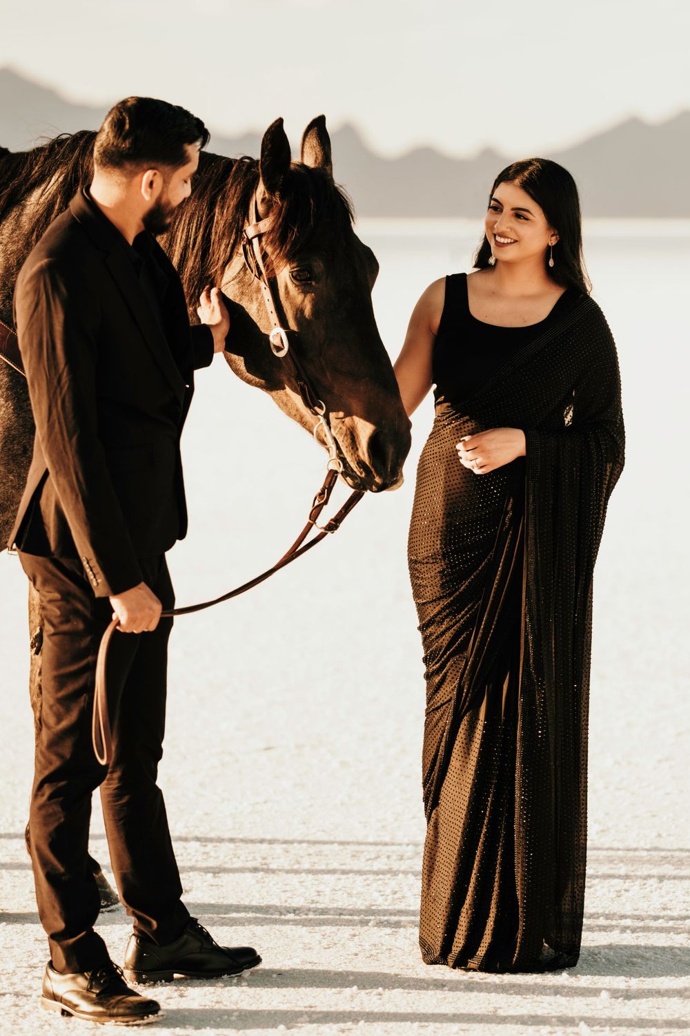 Couple in formal attire poses with a brown horse on a salt flat during golden hour for an engagement photoshoot.