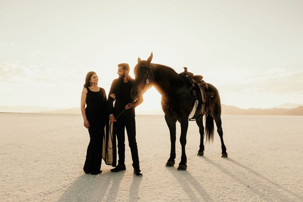 Two people in black outfits stand with their horses on a vast desert landscape at sunset.