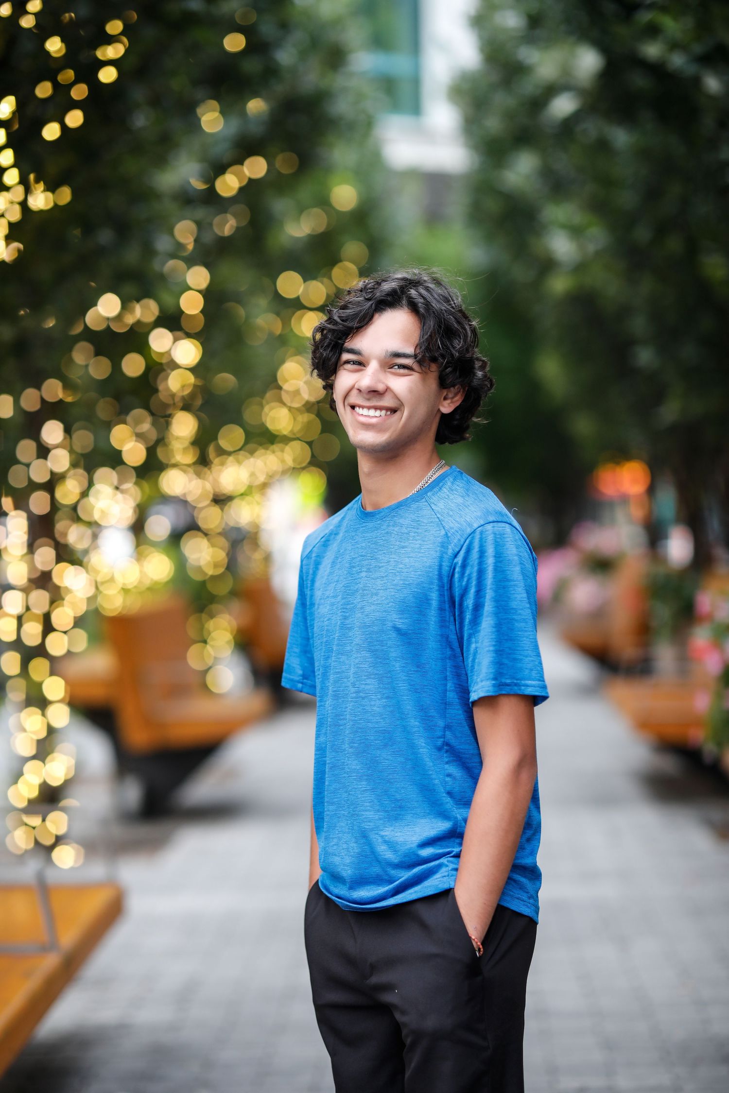 Smiling young man in blue t-shirt standing on urban walkway with bokeh string lights.