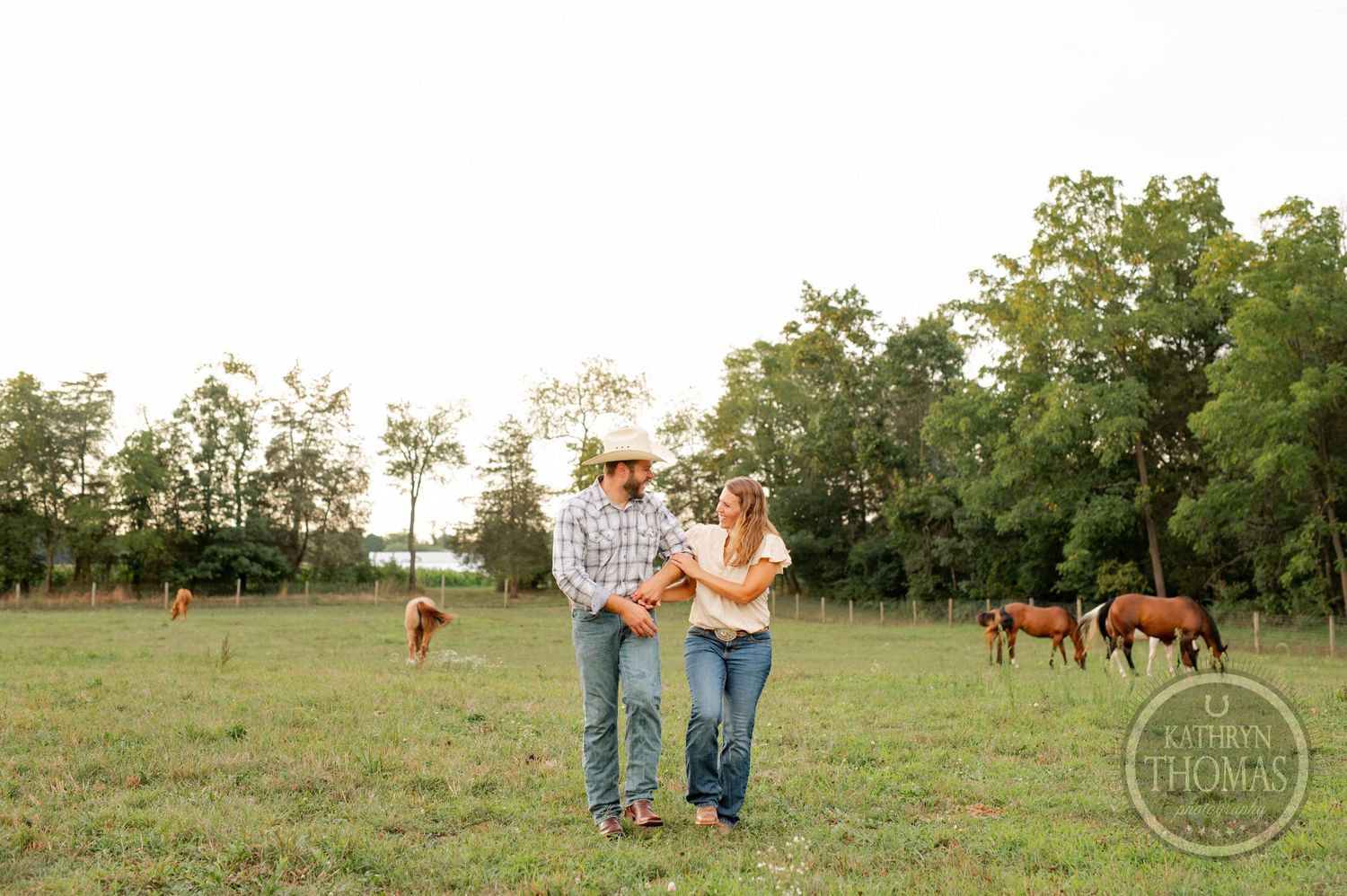 Couple standing together in pastoral field with horses grazing in background during golden hour.