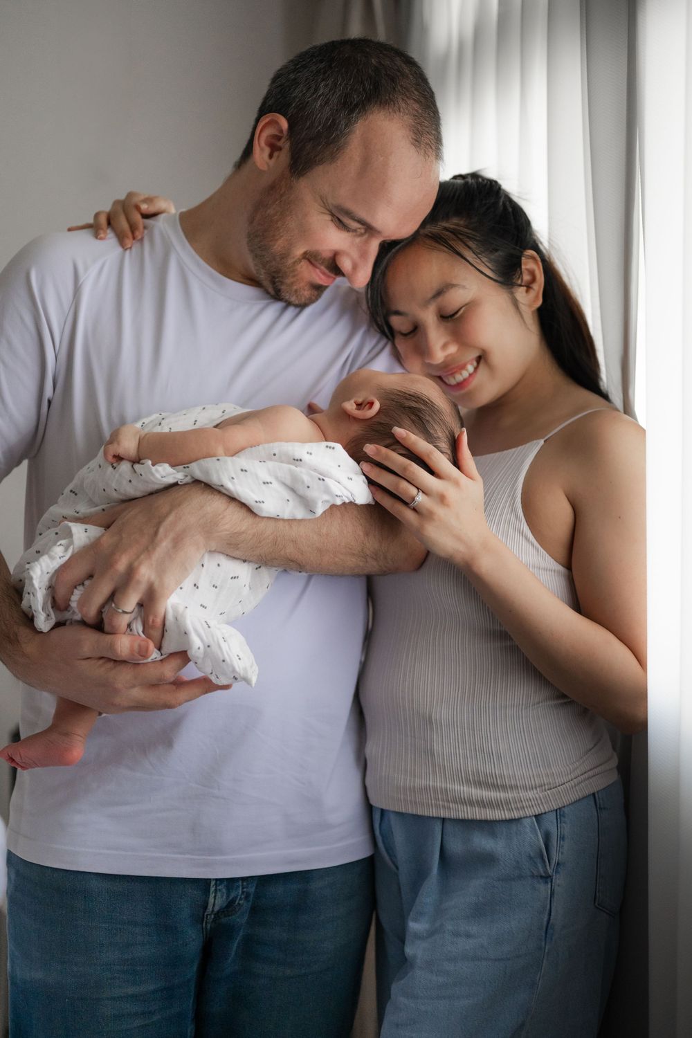 Mother and father cuddling their newborn by a bright window during a Phoebe Lee Photo session in Toronto